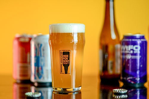 Glass full of golden ale with independent craft brewer seal and packaged beer in the background